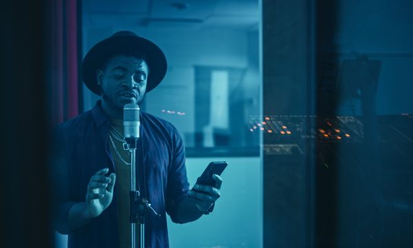 Portrait of Successful Young Black Artist, Singer, Performer Sin Portrait of Successful Young Black Artist, Singer, Performer Singing His Hit Song for the New Album. Wearing Stylish Hat, Holding Smartphone and Standing in Music Record Studio Soundproof Room.